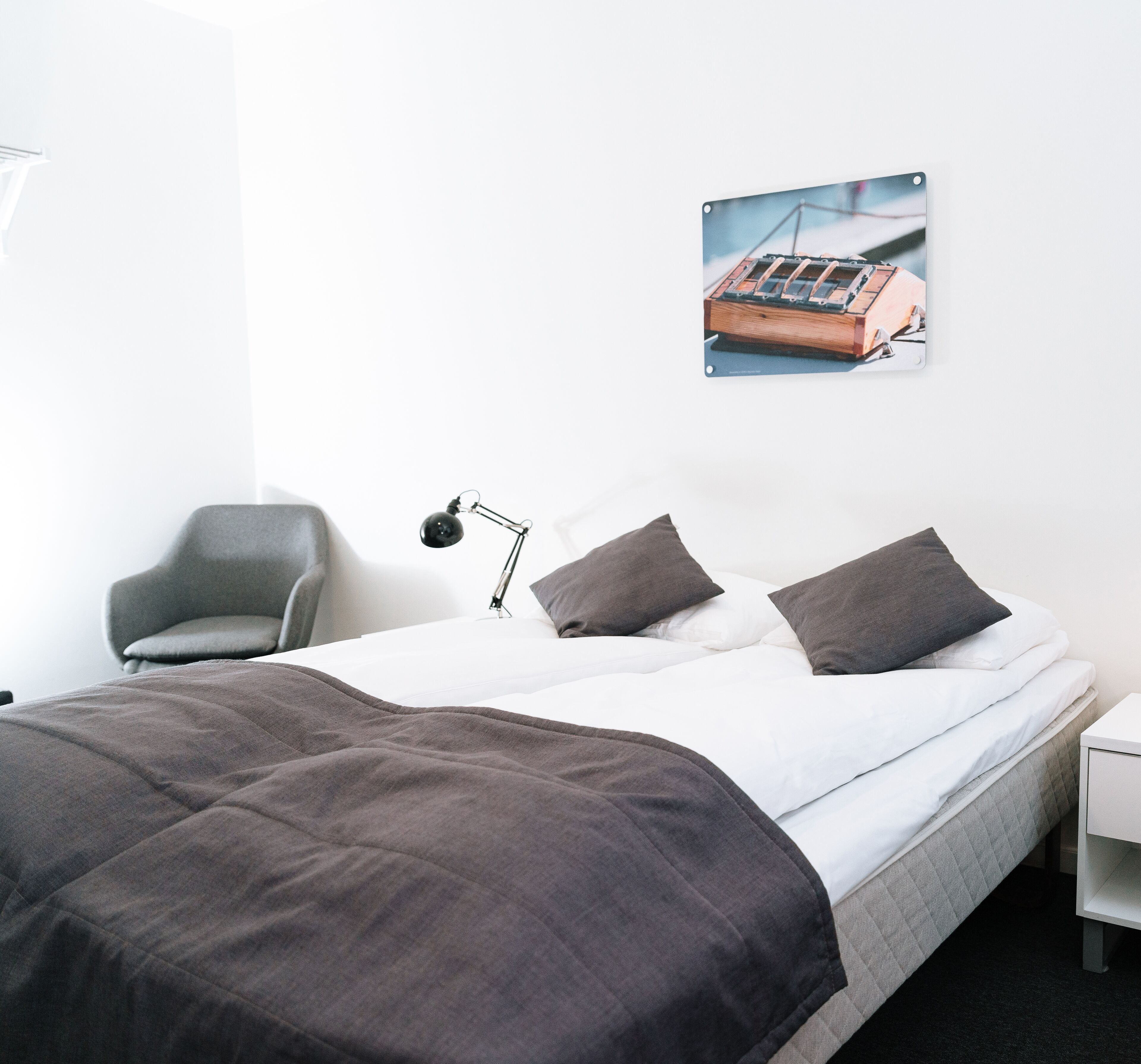 A neatly made bed with a gray comforter and pillows, a gray chair, a desk with a lamp, and a wall-mounted picture of a train.