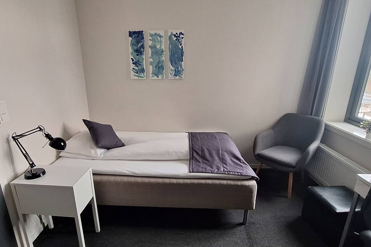 A small, neatly arranged bedroom with a bed, desk, chair, and a window with a view.