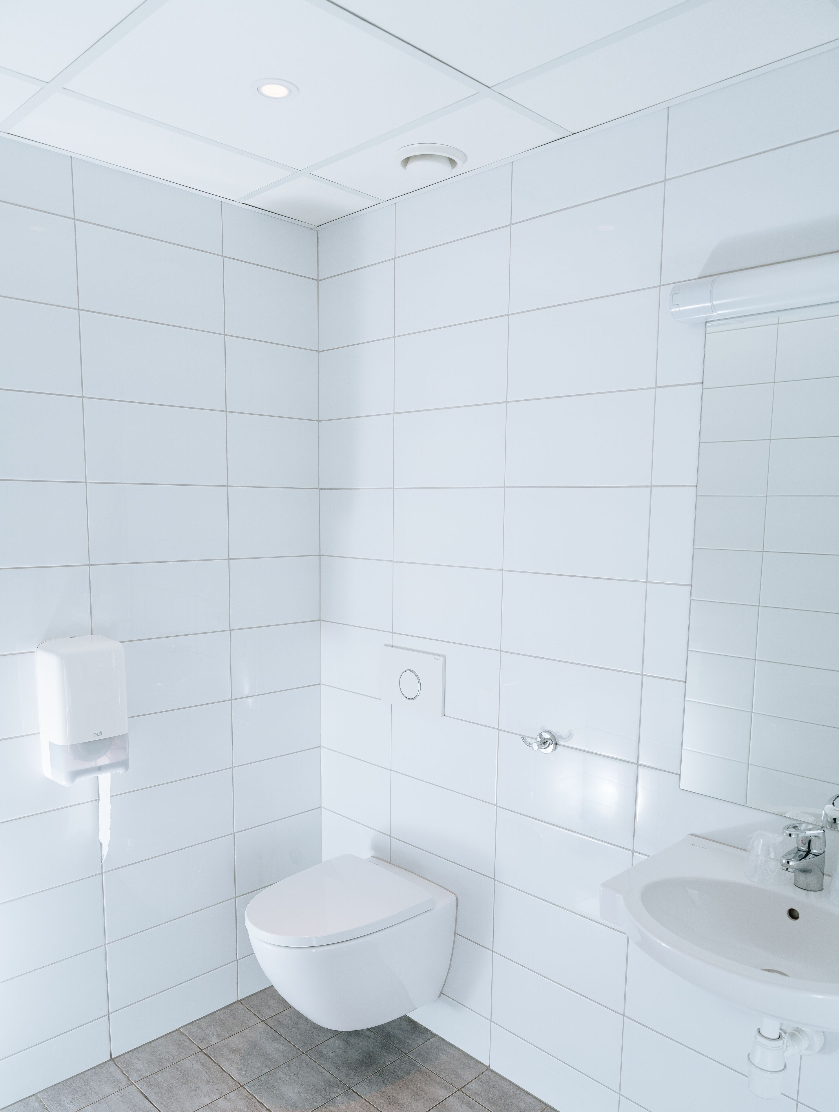 Moderni kylpyhuone, jossa on valkoiset laatat, wc-istuin, pesuallas ja käsienkuivaaja.