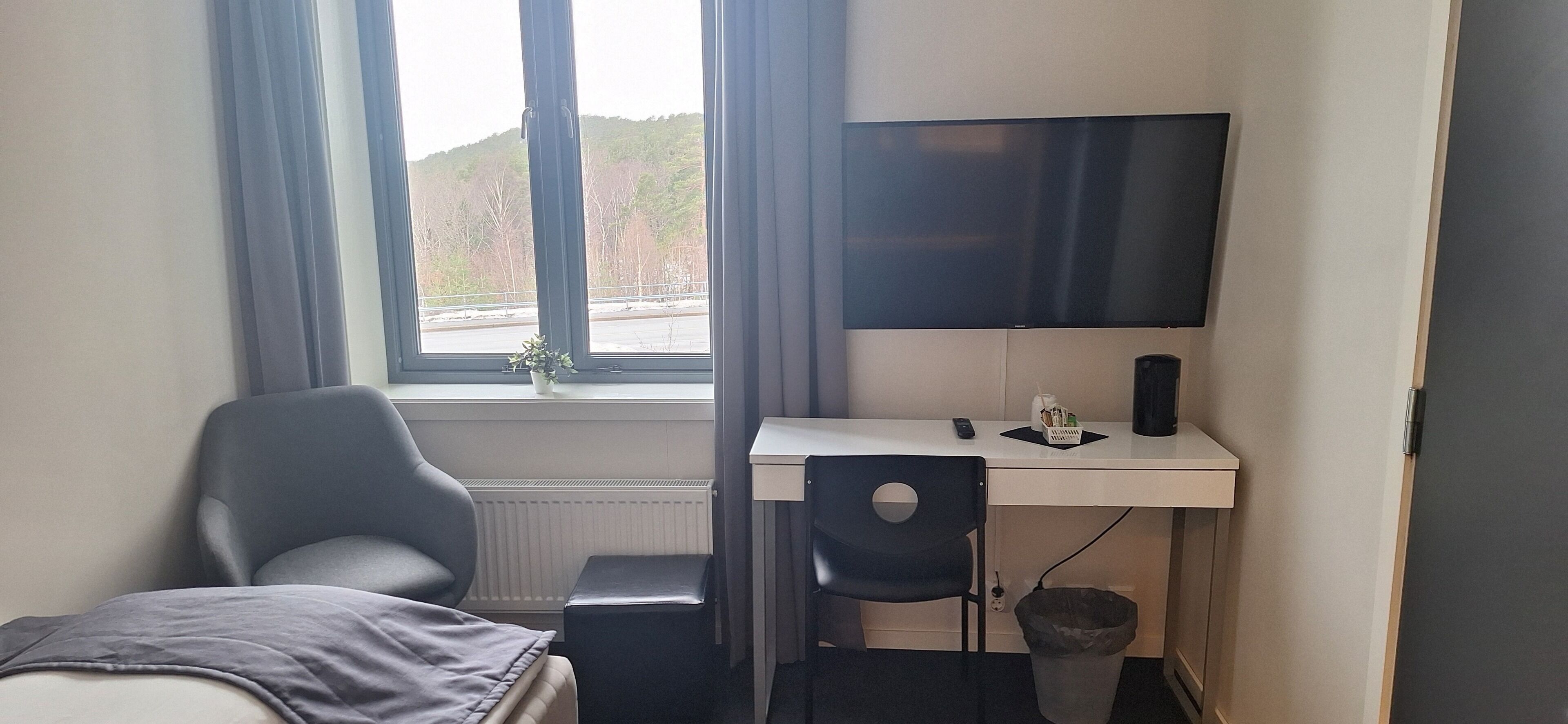 Een kleine, moderne kamer met een bed, een bureau, een stoel, een televisie en een raam met uitzicht op bomen en heuvels.