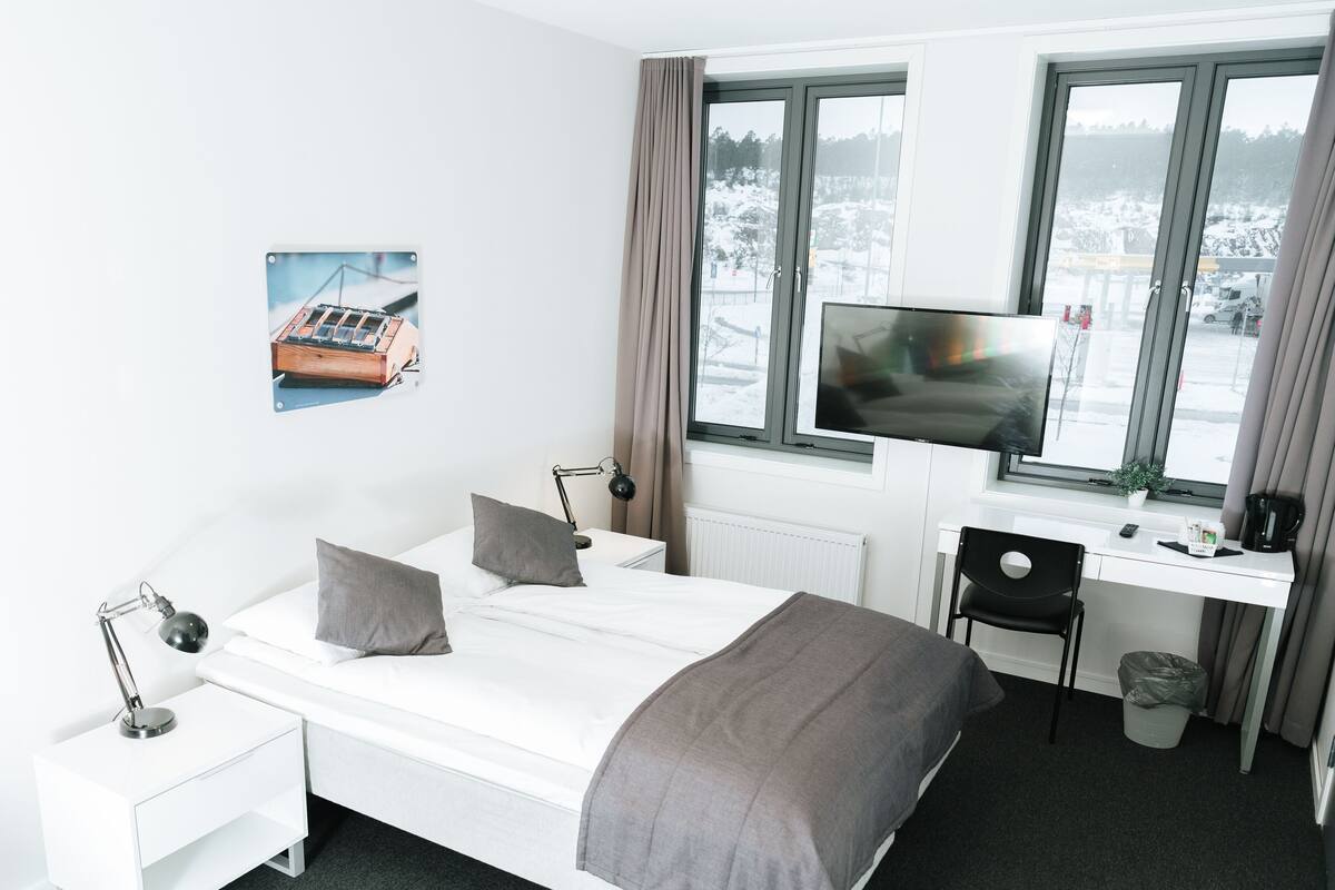 A modern hotel room with a bed, desk, chair, TV, and a window with a view of a snowy landscape.