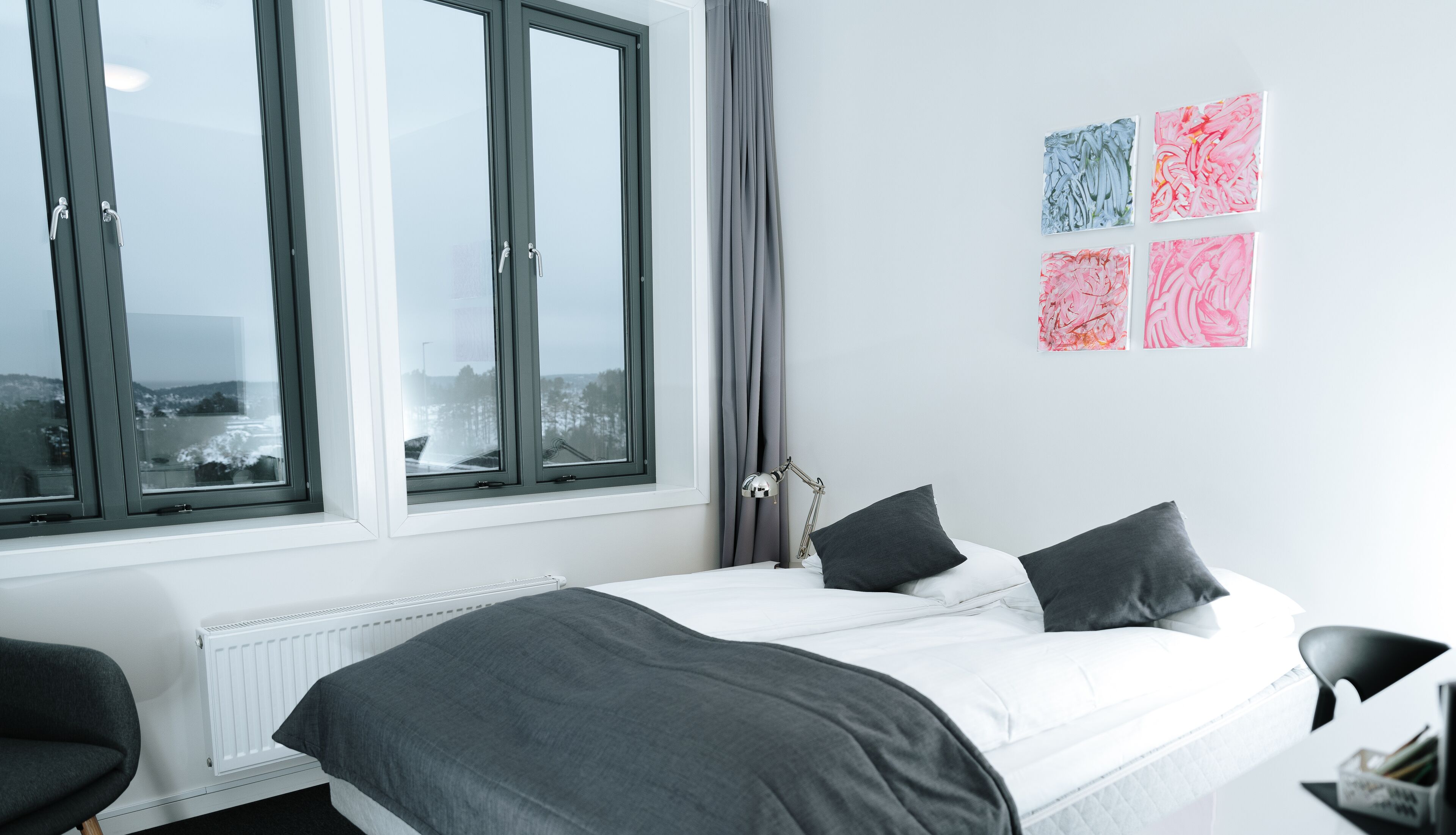 A modern bedroom with a large window, a bed with dark bedding, a desk, and a chair.