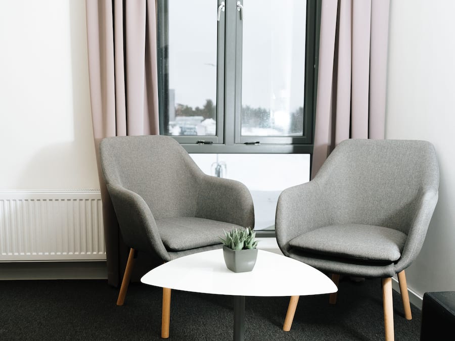 A modern room with two grey armchairs, a white table, and a window with curtains.