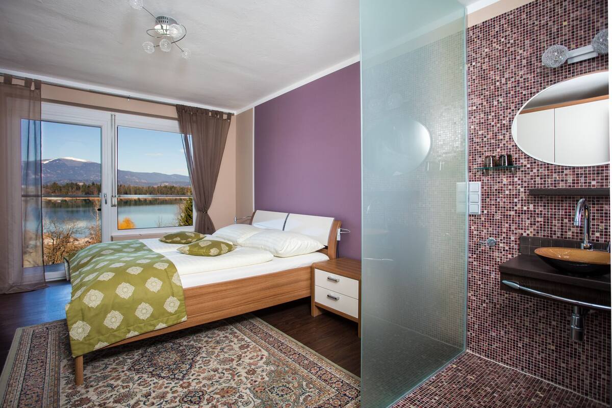Una camera da letto con un letto, un comodino, una finestra vista lago e montagne, e un bagno con un lavandino e uno specchio.