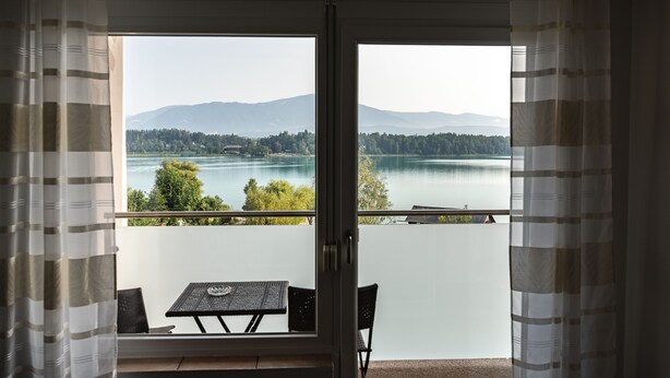 A balcony with a table and chairs, overlooking a lake with mountains in the background. - Faaker See Inn (Finkenstein am Faaker See)