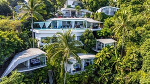 Une maison moderne à plusieurs étages, entourée d’une végétation luxuriante, dotée d’un grand balcon en verre et de plusieurs niveaux avec des espaces de détente en plein air.