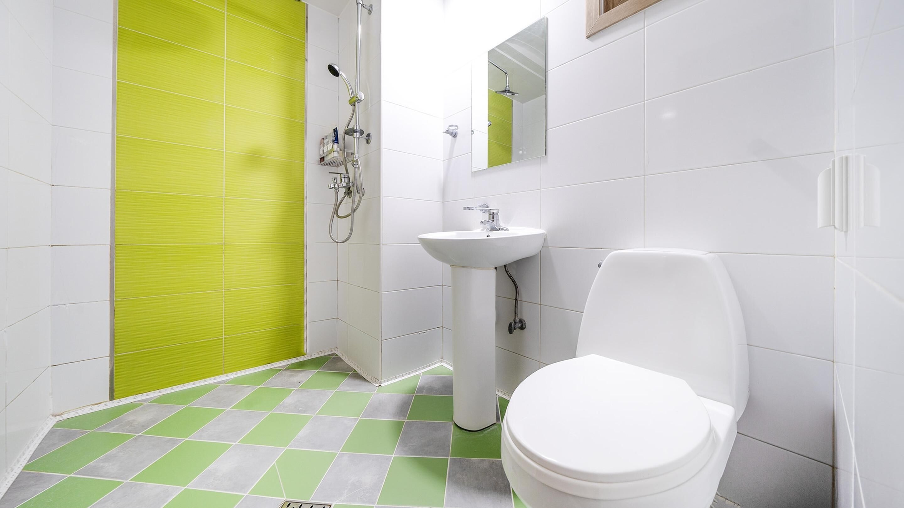 Casa de banho com sanita branca, lavatório e chuveiro. O chão tem um padrão xadrez verde e branco.
