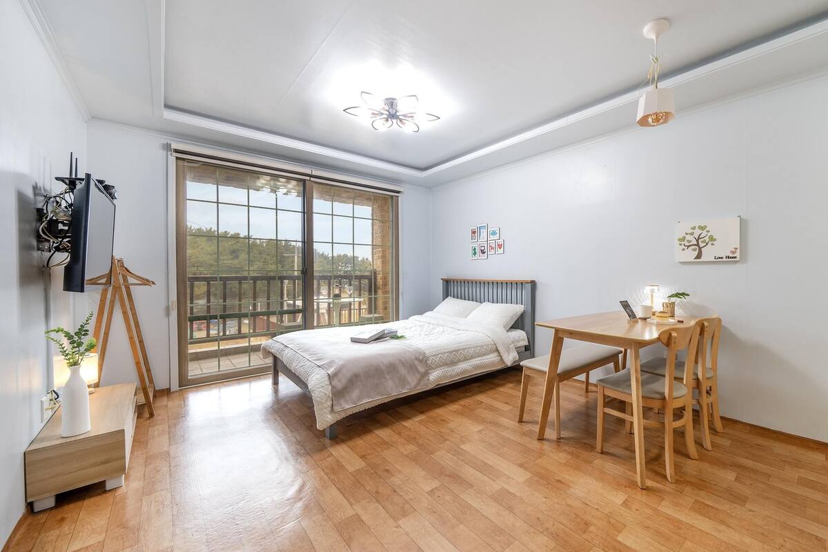 A bedroom with a bed, a desk, and a chair. There is a window with a view of greenery outside.