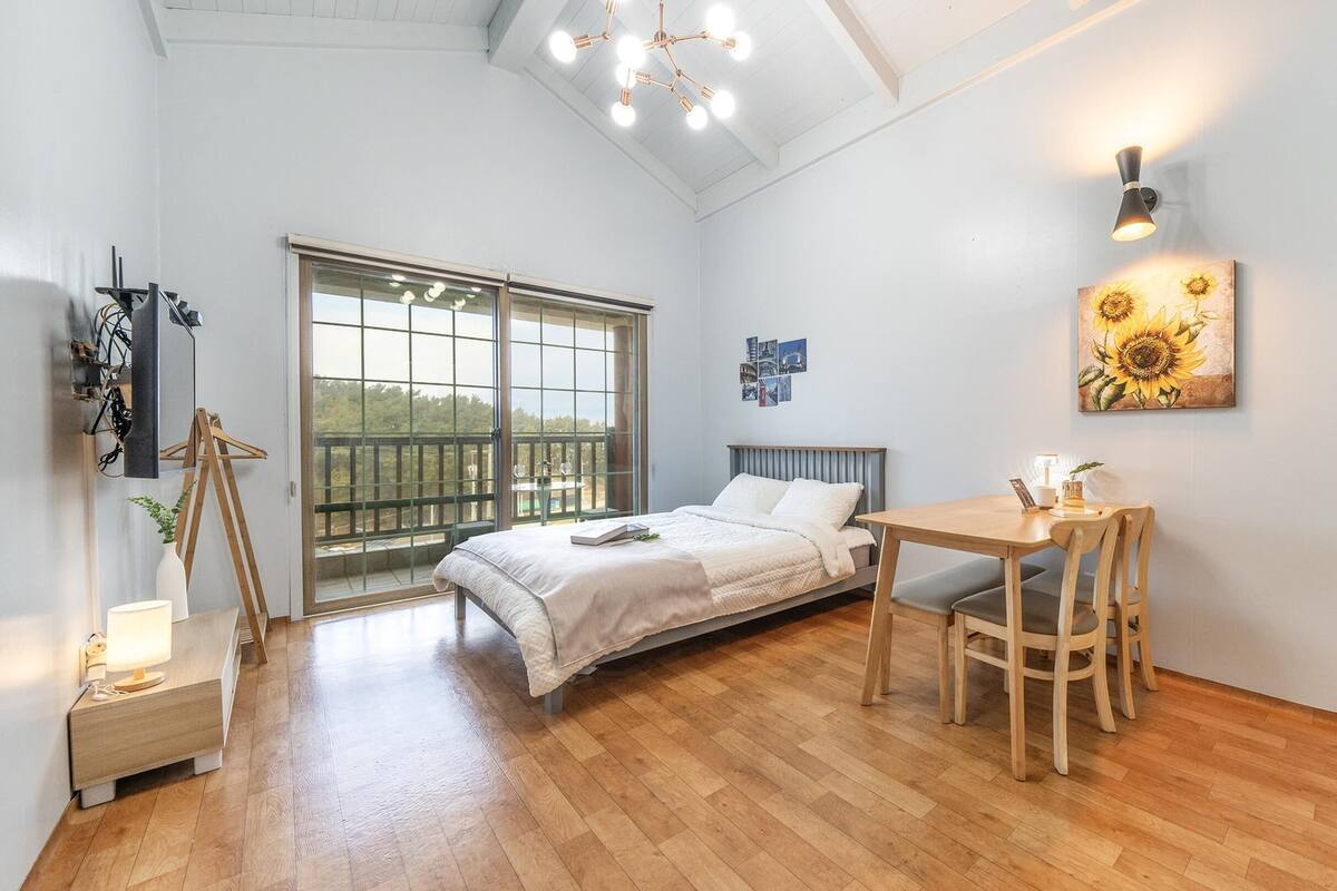 A bedroom with a bed, a desk, and a chair. There is a window with a view of trees.