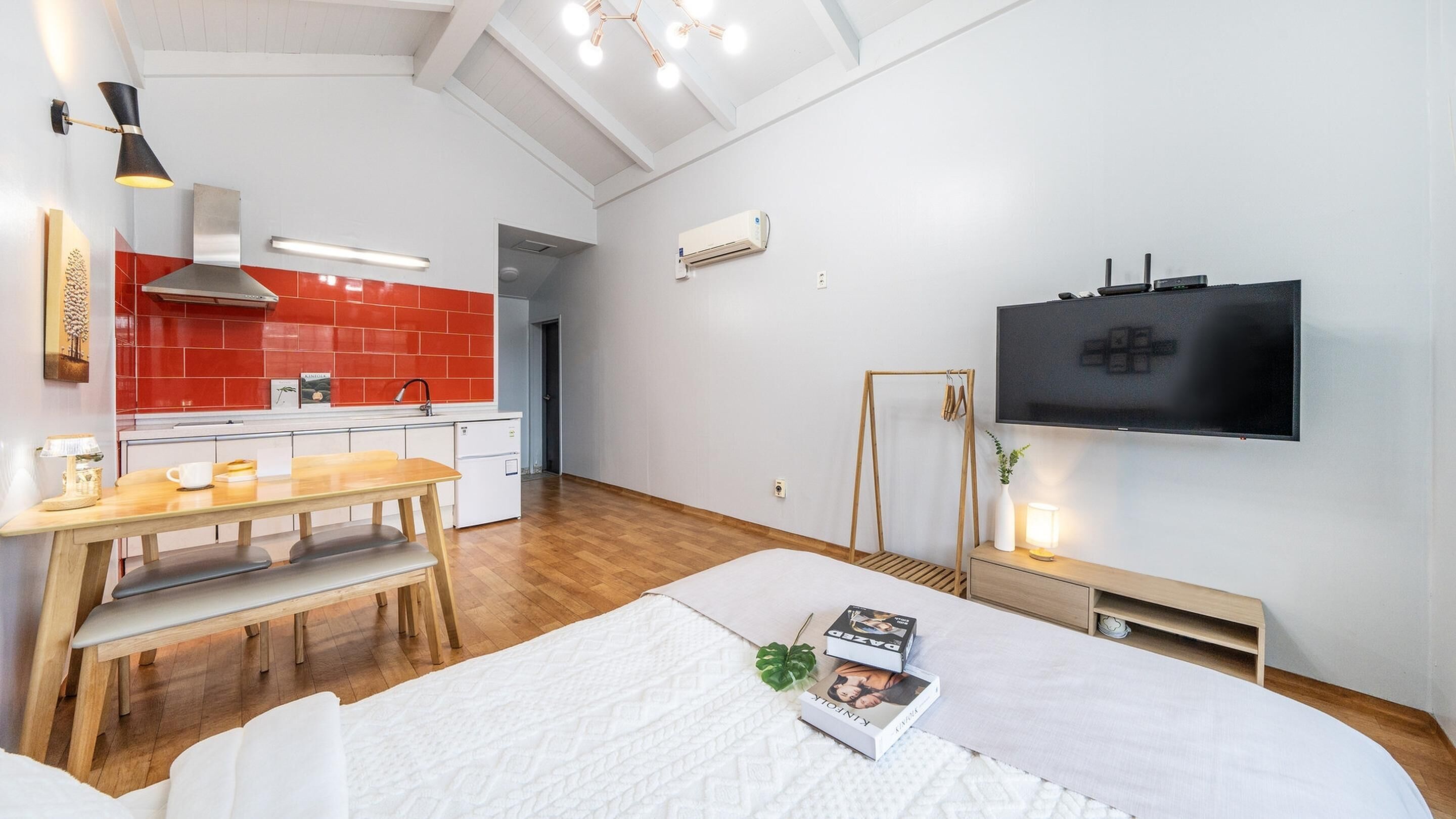 Una habitación de hotel moderna con piso de madera, una cama blanca, una mesa de comedor con sillas, una cocina con baldosas rojas, un televisor y una planta.