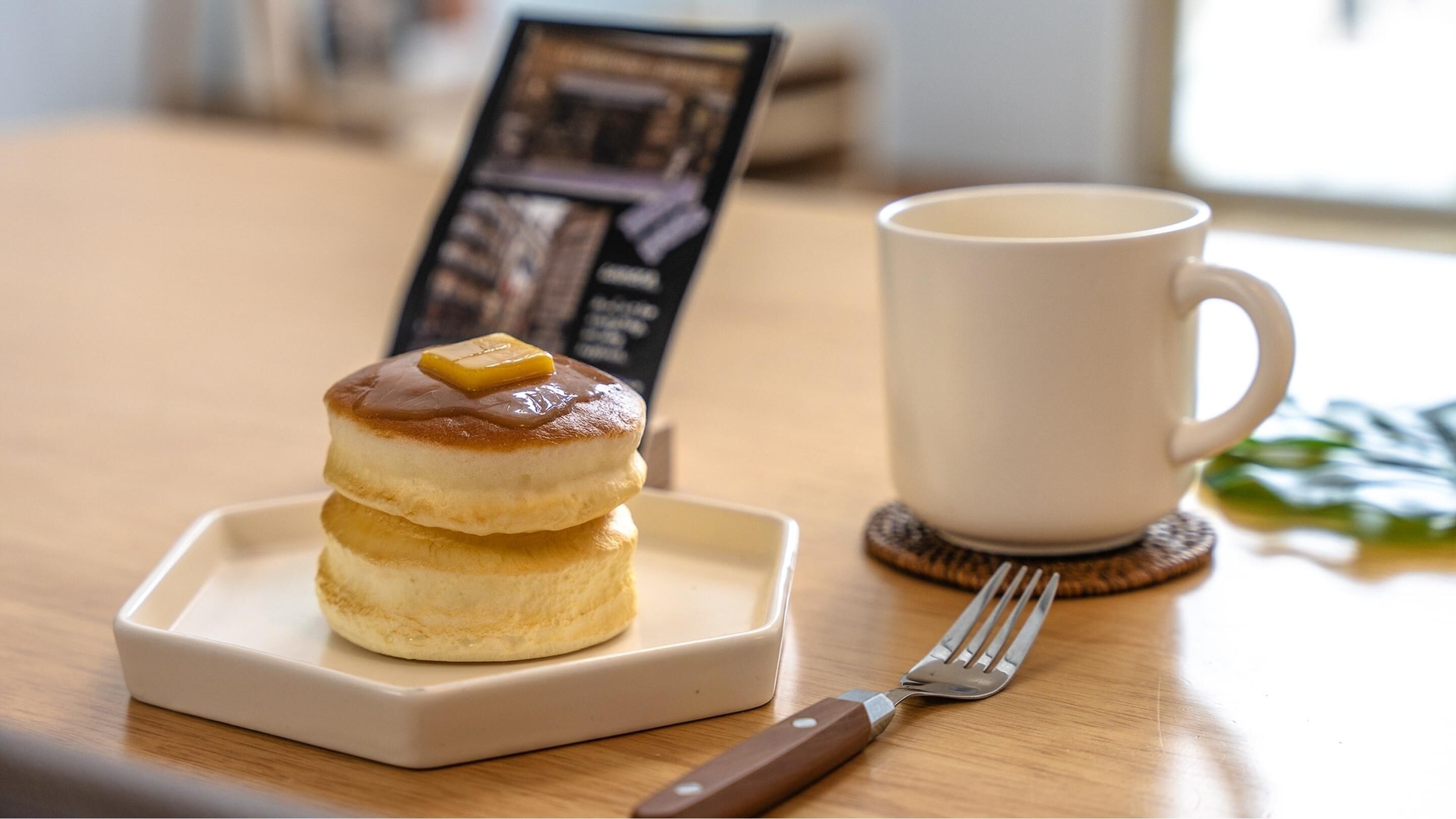 A stack of pancakes on a square plate, a white mug, and a fork on a wooden table.