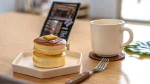 Una pila de tortitas sobre un plato cuadrado, una taza blanca y un tenedor en una mesa de madera.