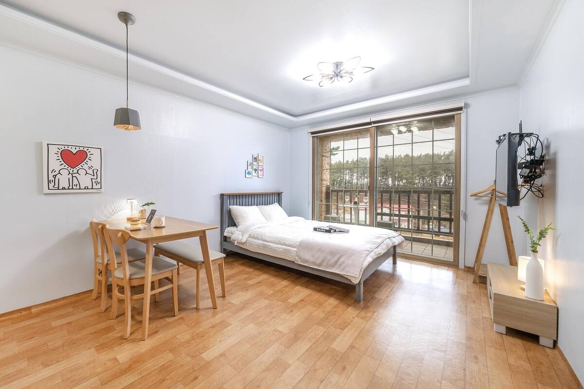 A bedroom with a bed, a desk, chairs, a ceiling fan, and a large window.