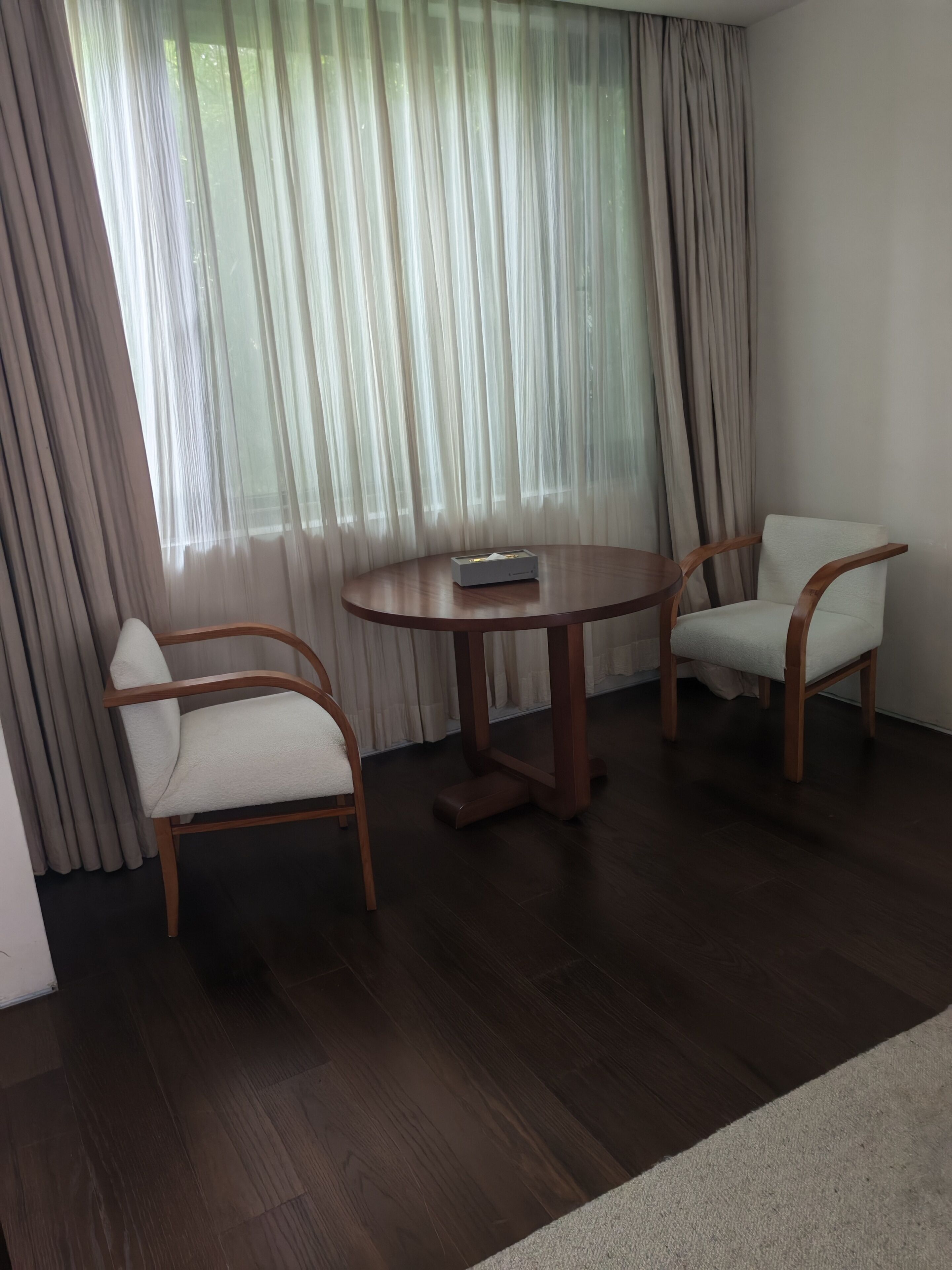 Una habitación con una mesa redonda de madera y dos sillas, un televisor sobre la mesa, y un ventanal con cortinas.