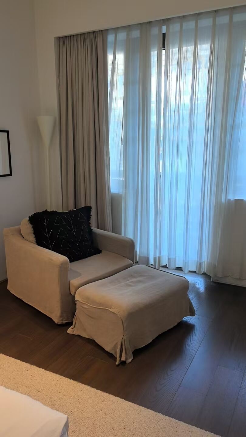 Un salon avec un canapé et un pouf, une lampe et une fenêtre ornée de rideaux légers.