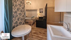 Ein kompaktes Hotelzimmer mit Bett, kleinem Tisch, Sofa, einer Kristallleuchtreiter, Waschbecken und einer Tür zu einem Badezimmer.