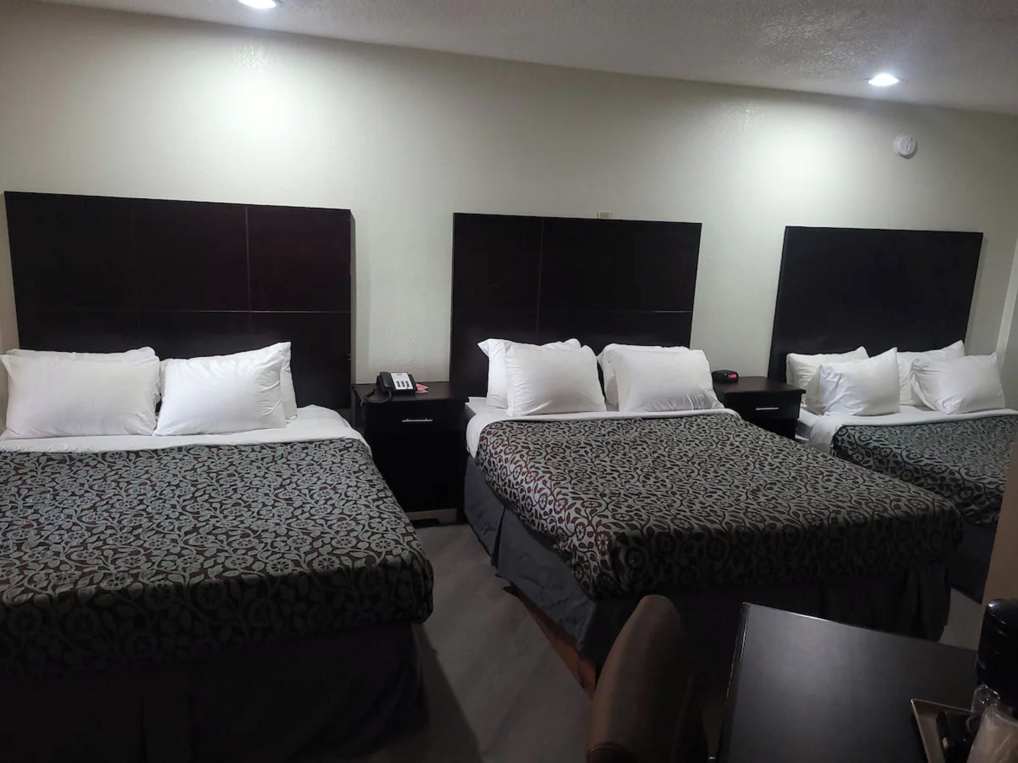 A hotel room with three beds, each with a patterned bedspread, a bedside table with a phone, and a wall-mounted light.