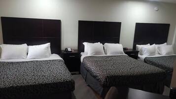 A hotel room with three beds, each with a patterned bedspread, a bedside table with a phone, and a wall-mounted light.