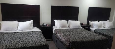 A hotel room with three beds, each with a patterned bedspread, a bedside table with a phone, and a wall-mounted light.