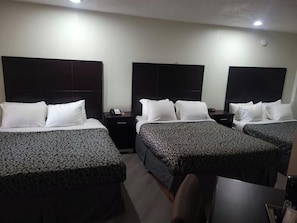 Ein Hotelzimmer mit drei Betten, jedes mit einer gemusterten Tagesdecke, einem Nachttisch mit Telefon und einer an der Wand montierten Lampe.