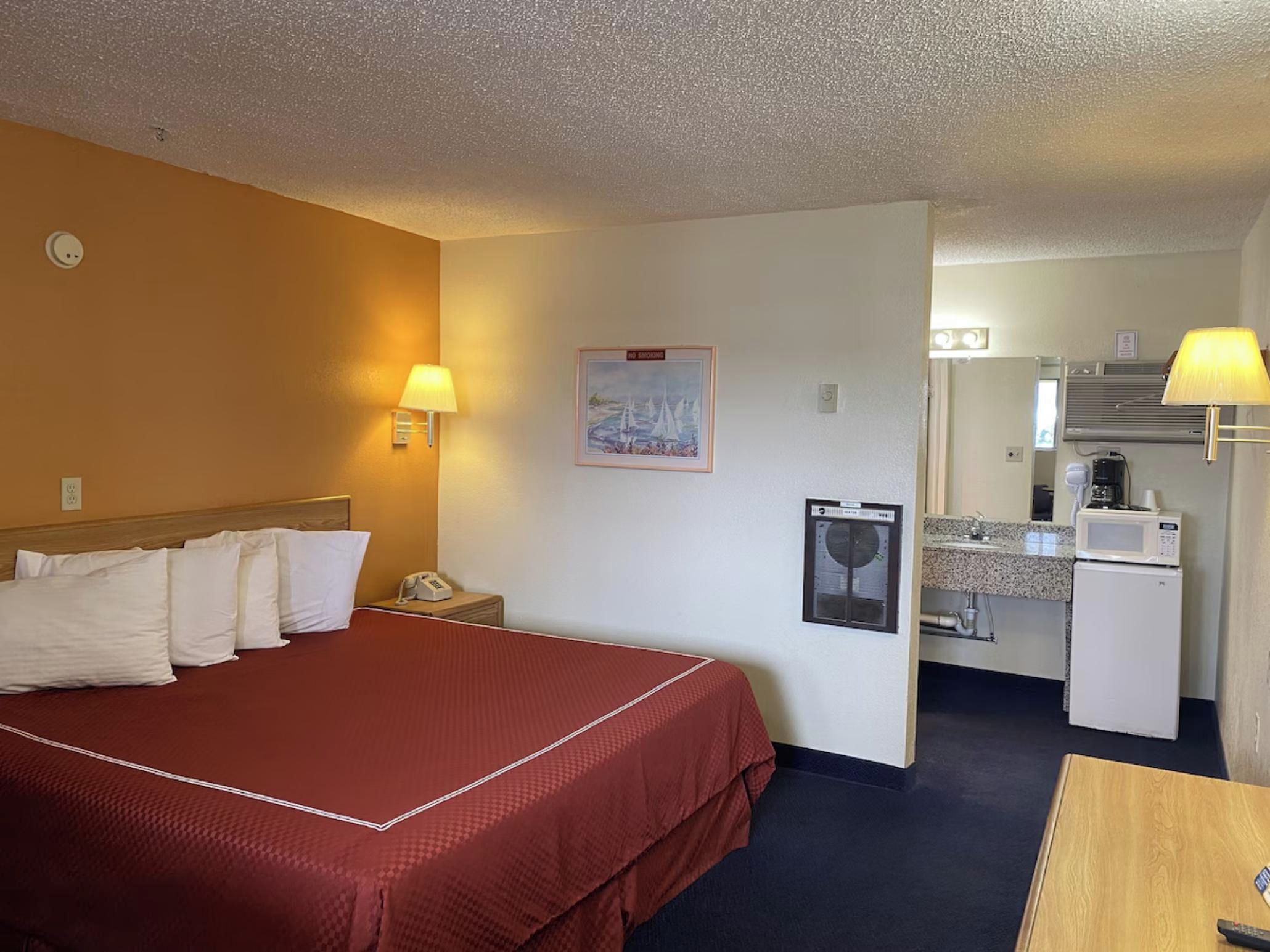 A hotel room with a bed, bedside lamps, a small desk with a microwave, and a painting on the wall.