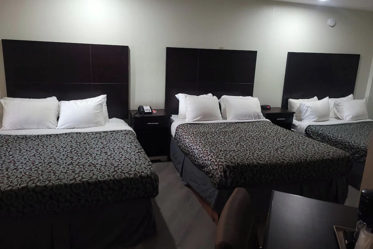 A hotel room with three beds, each with a patterned bedspread, a bedside table with a phone, and a wall-mounted light.