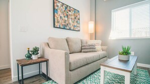 Ein Wohnzimmer mit einem beigen Sofa, einem gemusterten Teppich, einem kleinen Tisch mit einer Pflanze und einem an der Wand hängenden abstrakten Kunstwerk.