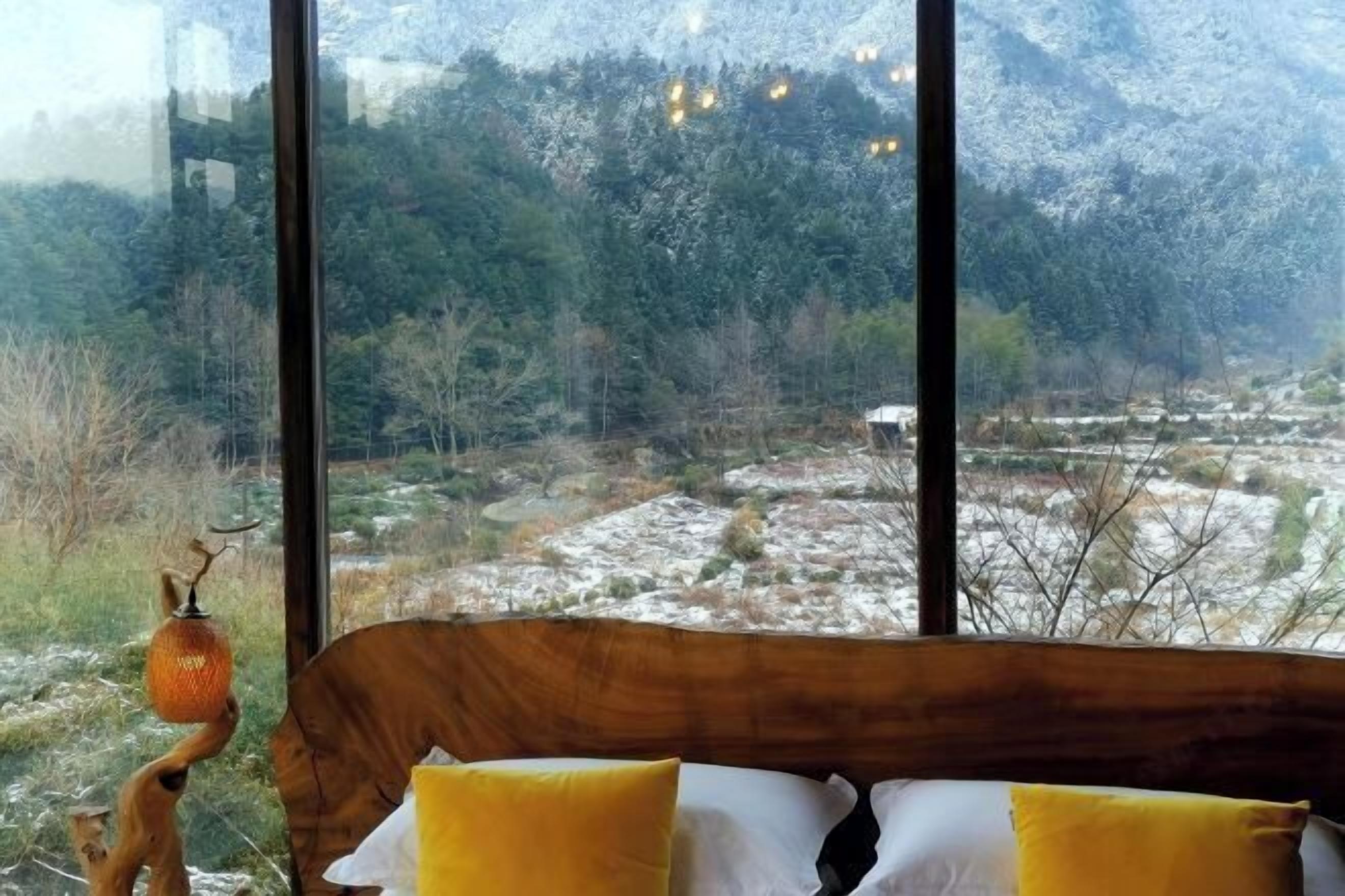 A room with a bed, a wooden headboard, and a view of a mountainous landscape through large windows.