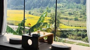 Ein Zimmer mit großem Fenster und Blick auf eine üppige Landschaft, einem kleinen Holztisch und einem Topfpflanzen.