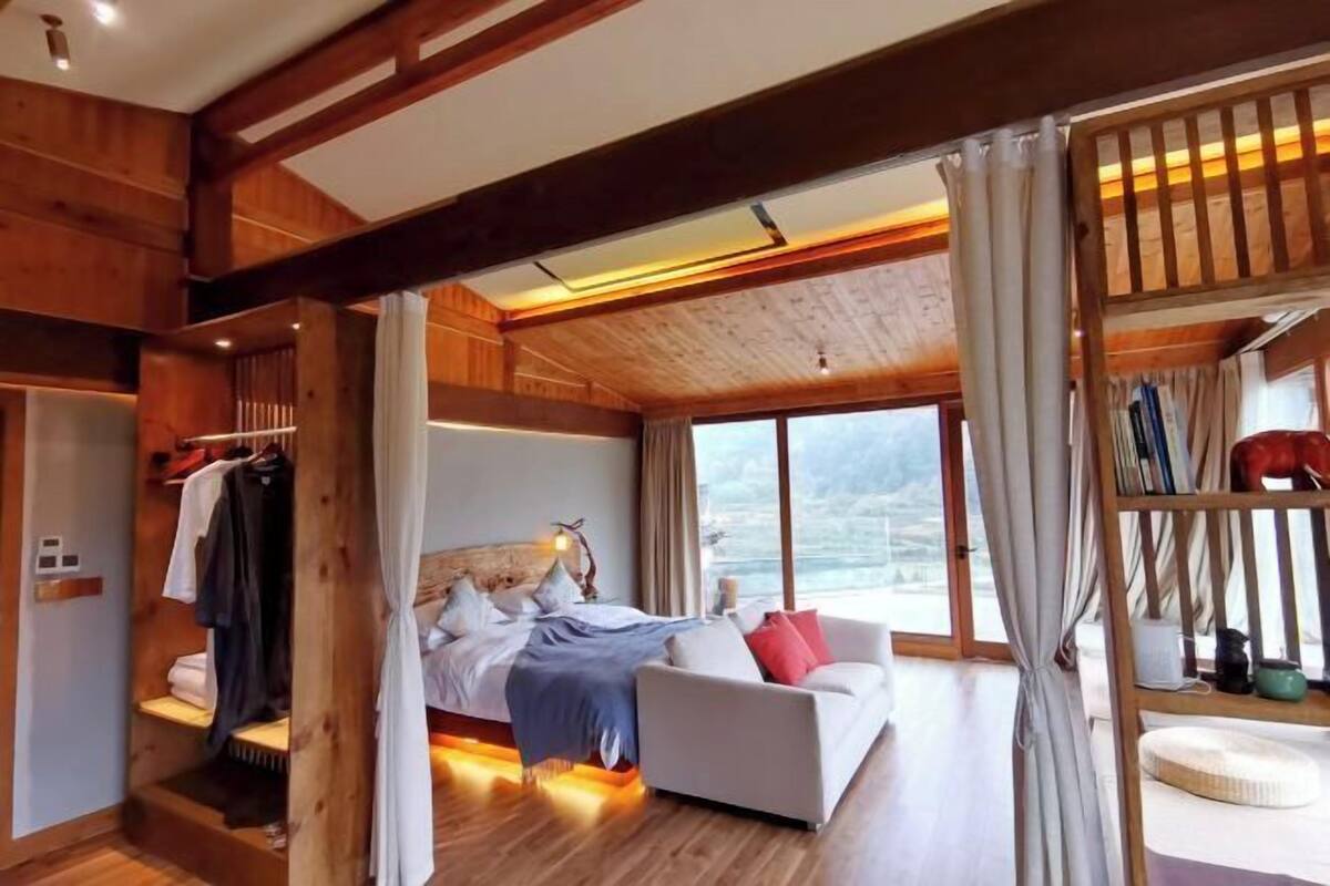 A bedroom with a bed, a white sofa, a wooden wardrobe, and a large window.