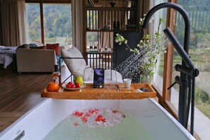 Une baignoire remplie de pétales de rose, un plateau avec des fruits et un vase, et une vue sur l’extérieur.