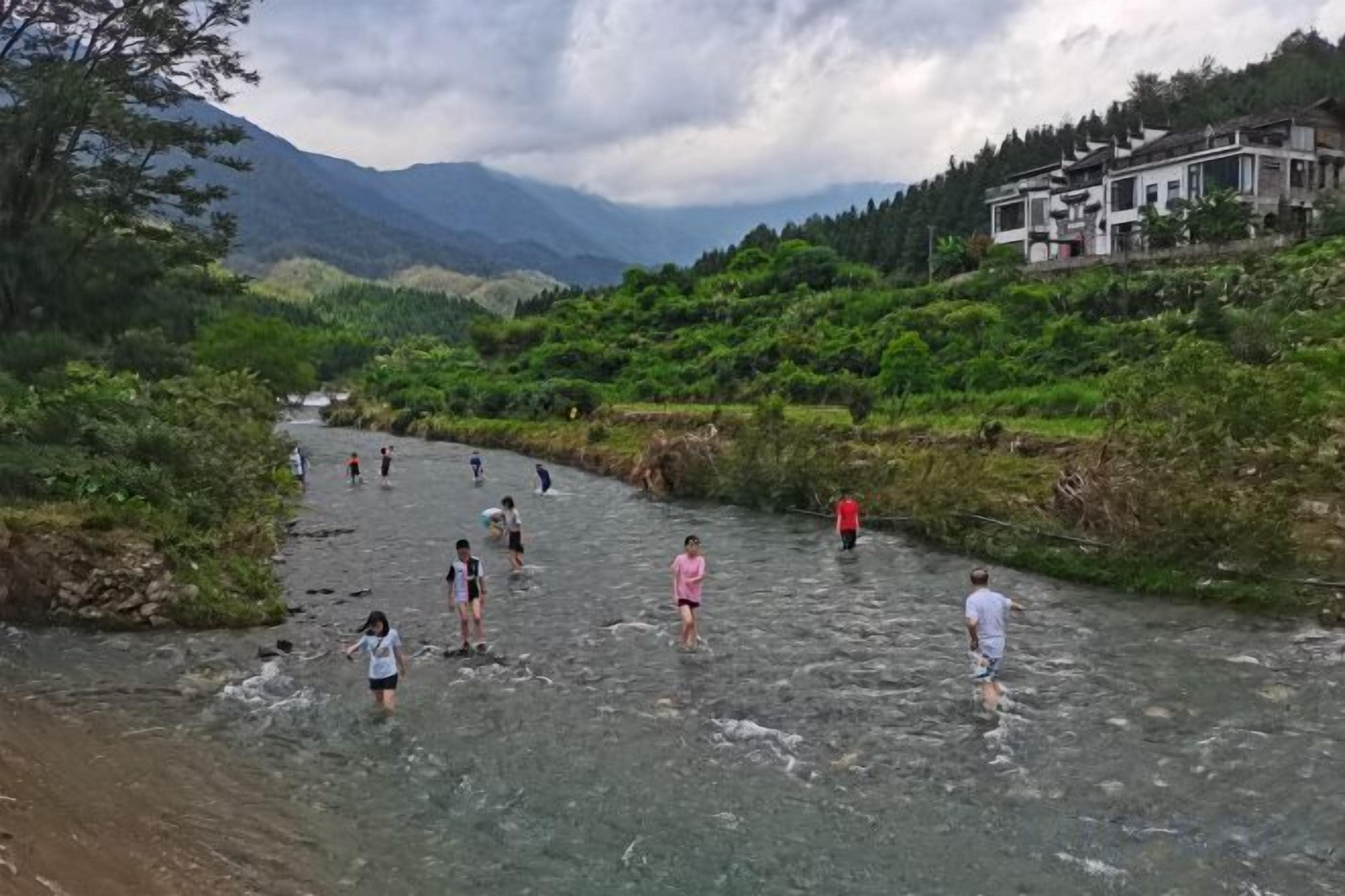 A river with people wading through it, surrounded by greenery and mountains in the background.