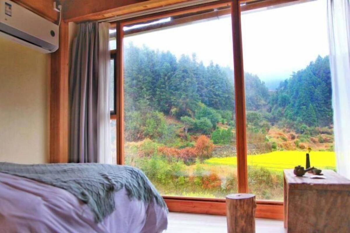 A bedroom with a large window, a bed, a wooden nightstand, and a view of a forested landscape.