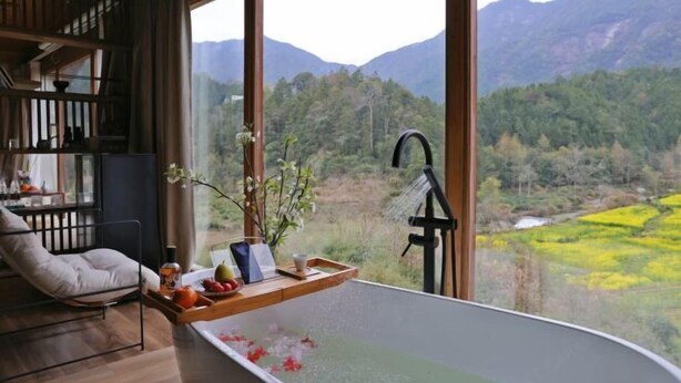 Ein Zimmer mit einem großen Fenster, einer Badewanne mit Blumen, einem Holzregal und einem Stuhl.