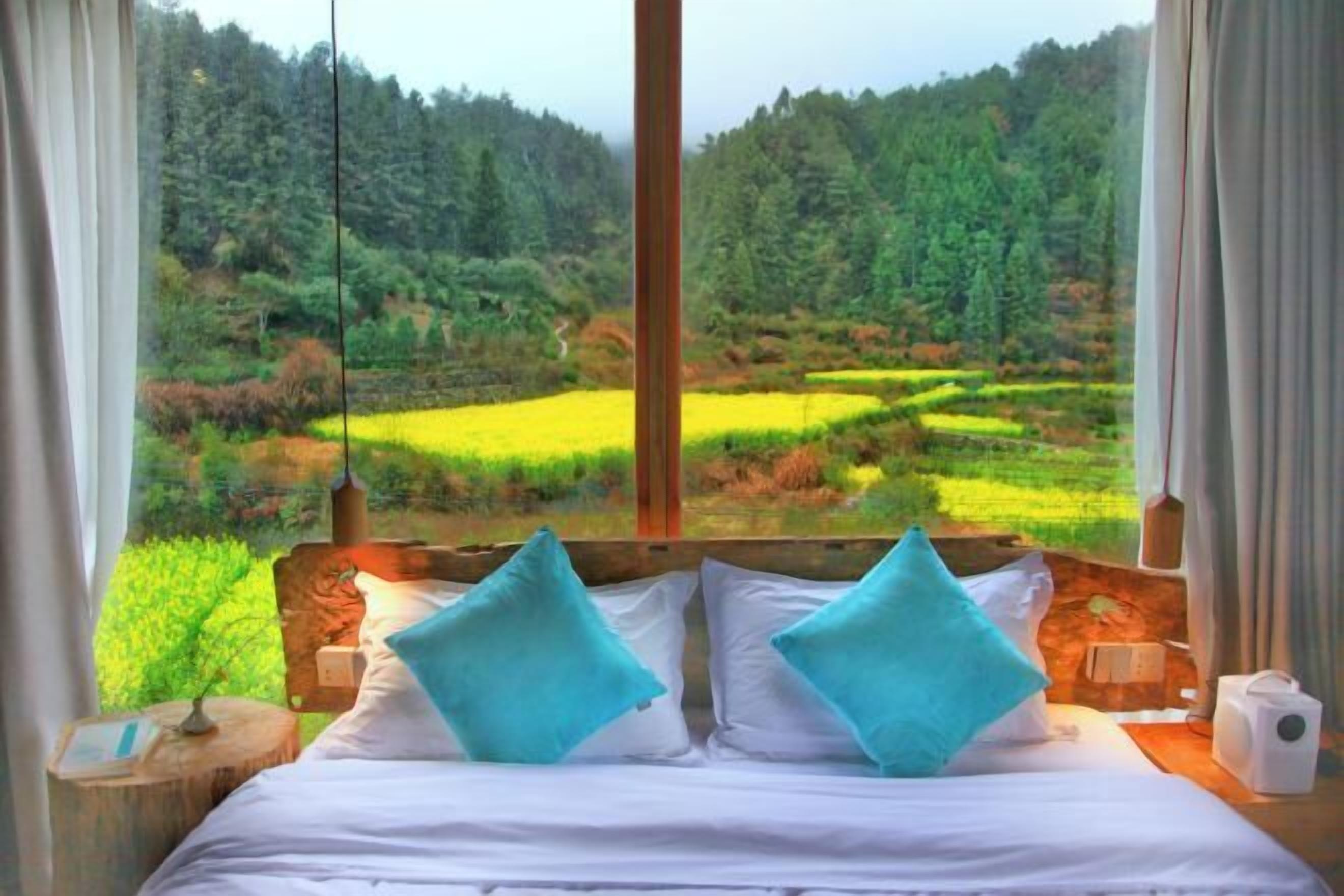 Una cama con sábanas blancas y dos almohadas azules, cabecera de madera, y vista a un paisaje boscoso a través de una ventana.