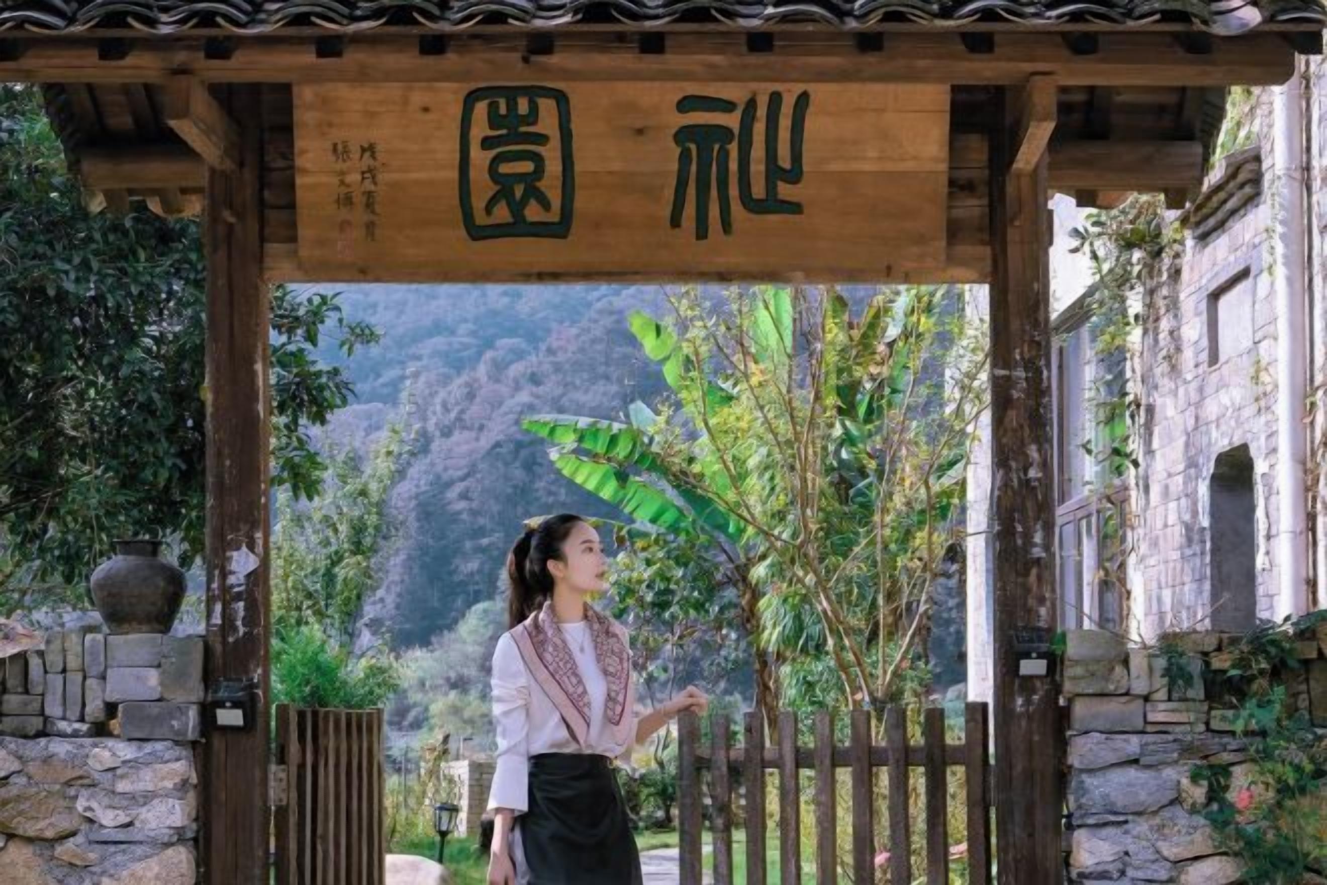 Un arco de madera tradicional con caracteres chinos, un muro de piedra y vistas a un paisaje montañoso.
