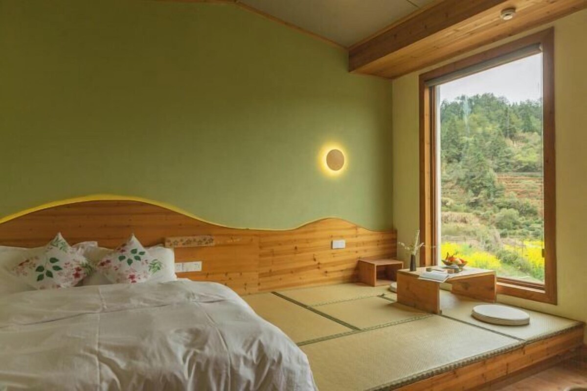 Un dormitorio con cabecera de madera, cama con ropa de cama blanca, mesita de noche pequeña, ventana con vistas a zonas verdes y una lámpara fijada a la pared.