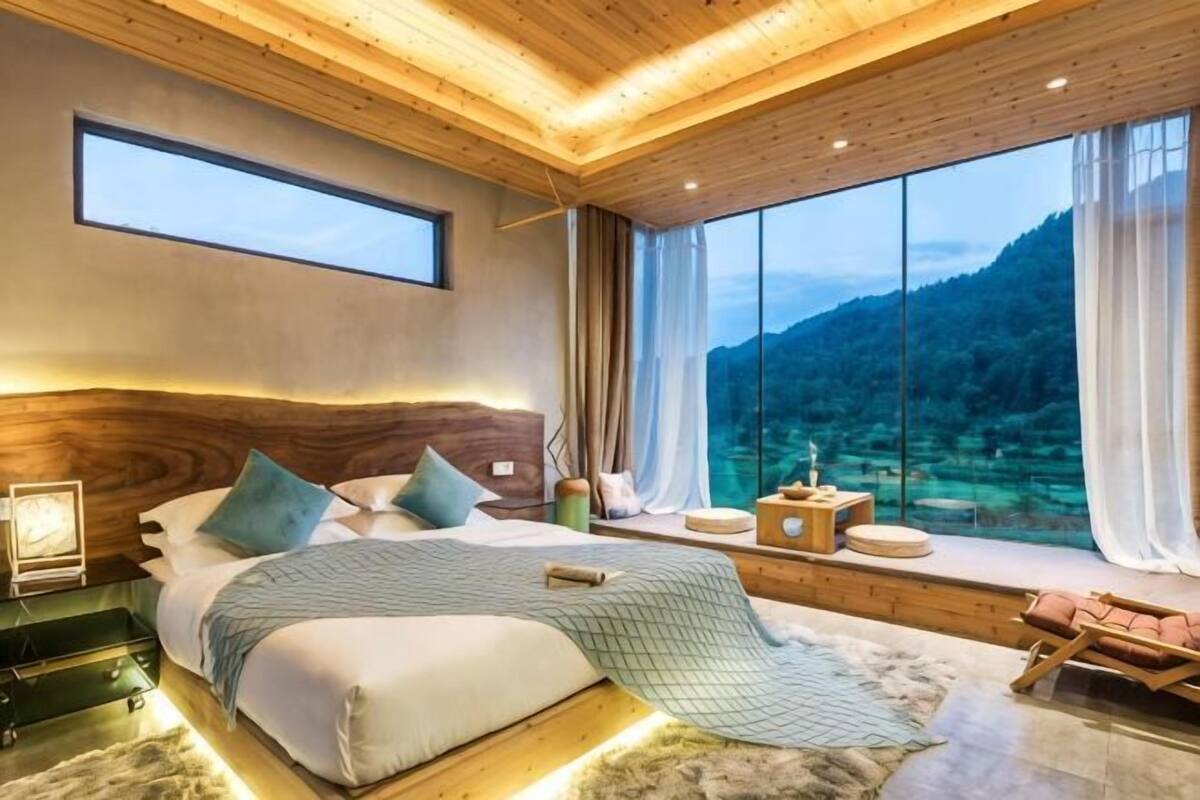 A bedroom with a large bed, a view of the mountains, a wooden ceiling, and a window with curtains.