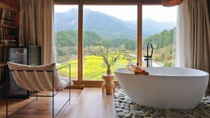 Ein modernes Badezimmer mit einer freistehenden Badewanne, einem Holzstuhl und Blick auf eine gebirgige Landschaft durch große Fenster.