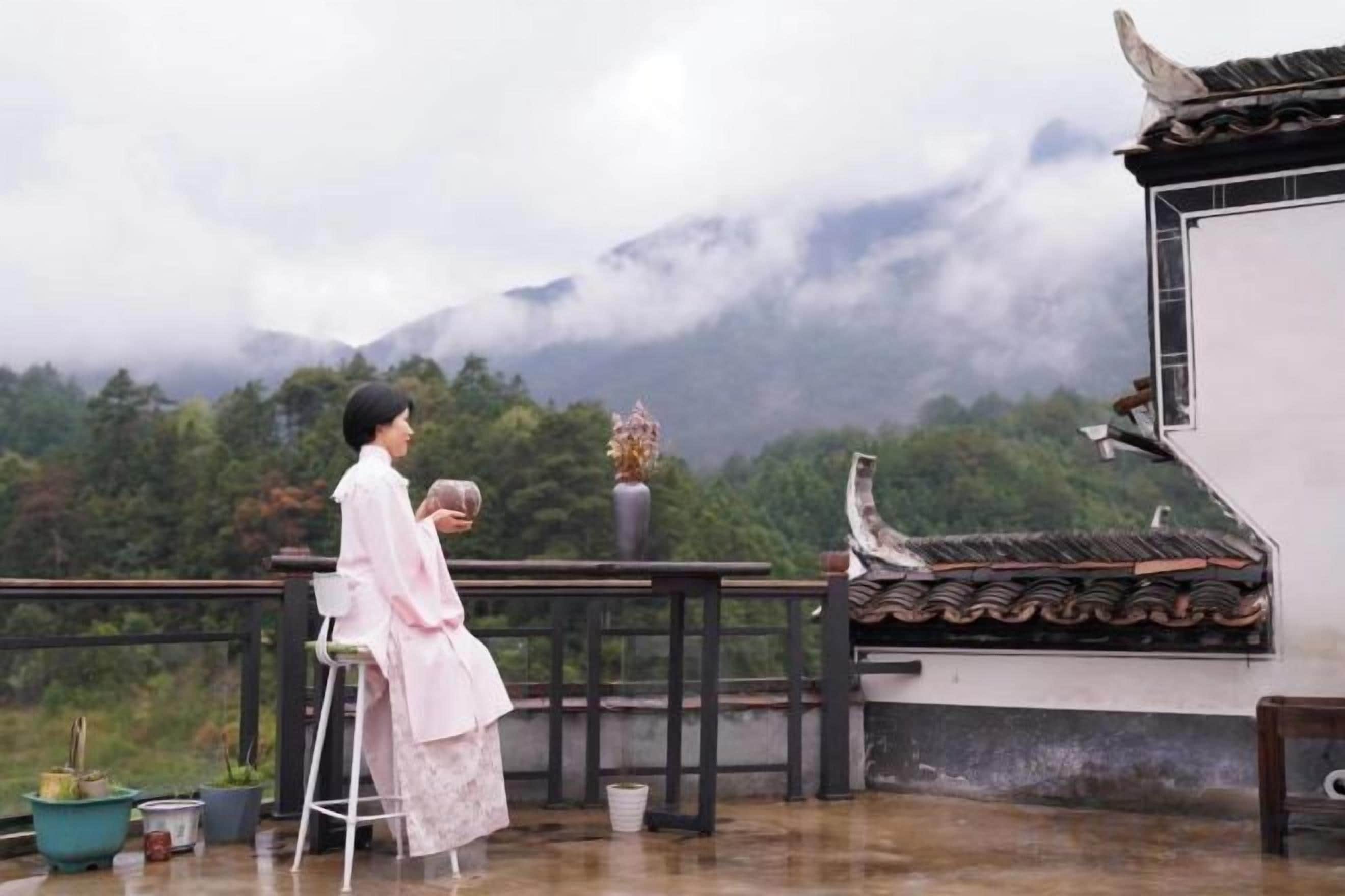 A person seated on a chair on a balcony overlooking a mountainous landscape with traditional architecture.