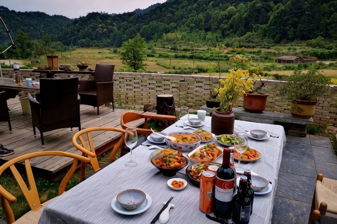 An outdoor dining area with a table set for a meal, surrounded by chairs, with a scenic view of a forested landscape.