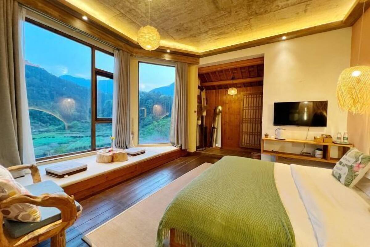 A bedroom with a large window, a bed, a TV, and a wooden ceiling.
