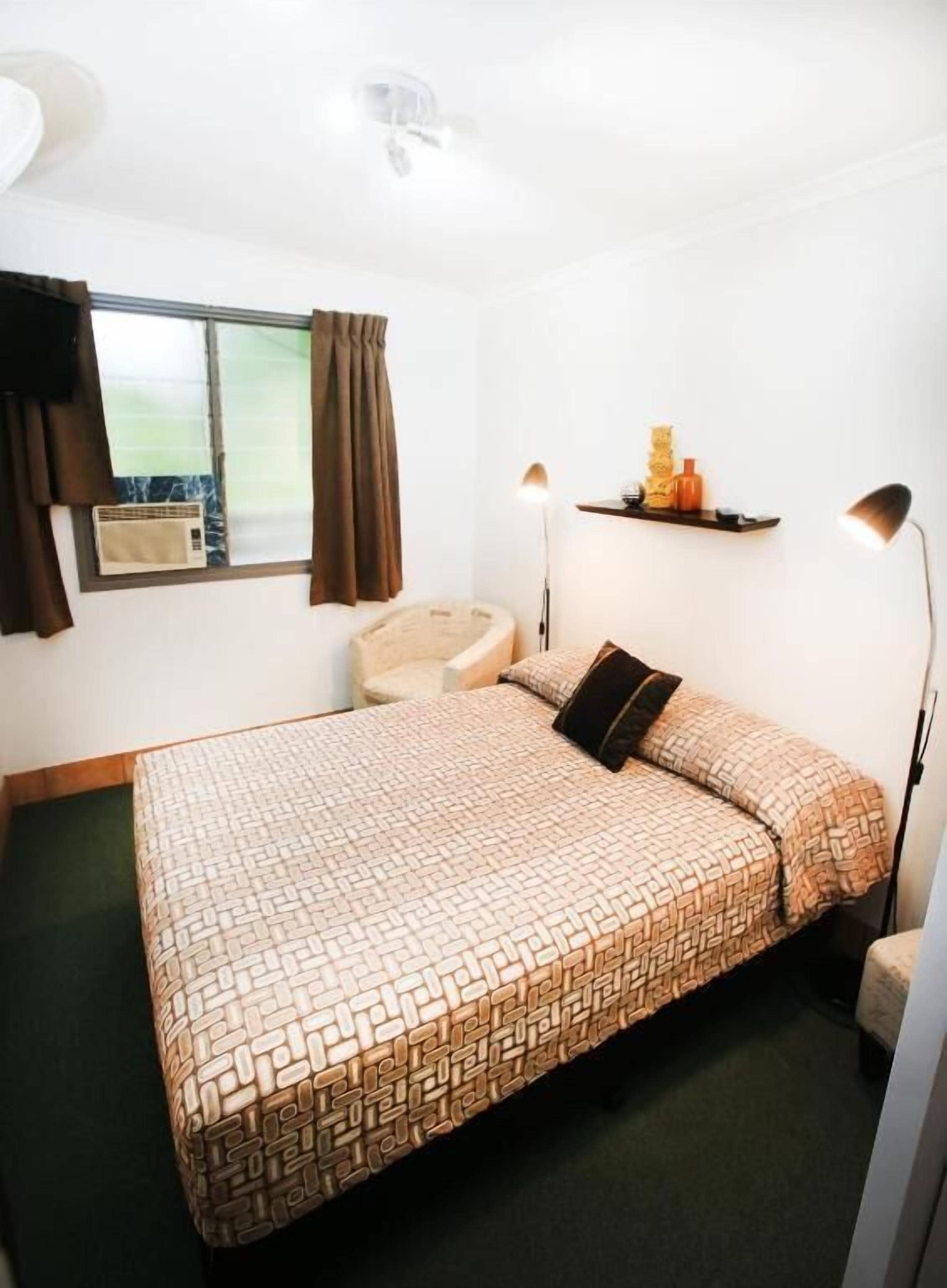 A single bed with a patterned bedspread, a bedside lamp, a small shelf with decorative items, and a window with curtains.