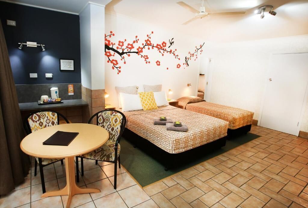 Habitación de hotel con dos camas, una mesita redonda pequeña, una silla, un escritorio con su silla y un estante en la pared.