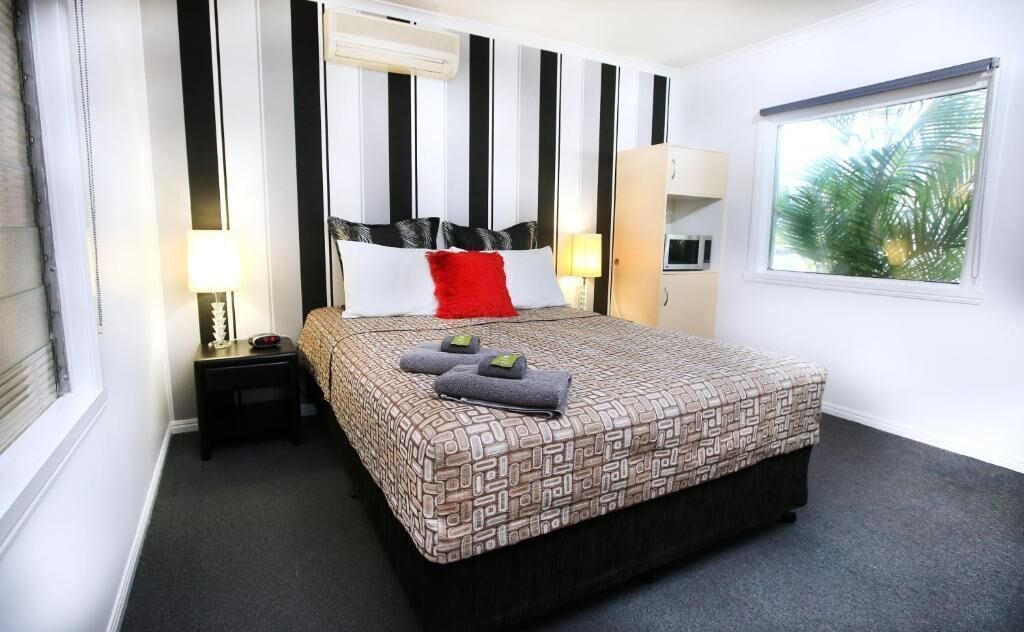 Una habitación de hotel con una cama, mesitas de noche, un ventanal y vistas a palmeras.