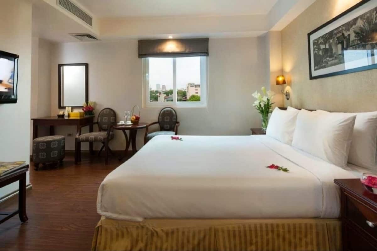 Ein Hotelzimmer mit einem großen Bett, einem Schreibtisch mit Stuhl, einem kleinen Tisch mit Lampe und einem Fenster mit Blick auf Gebäude.
