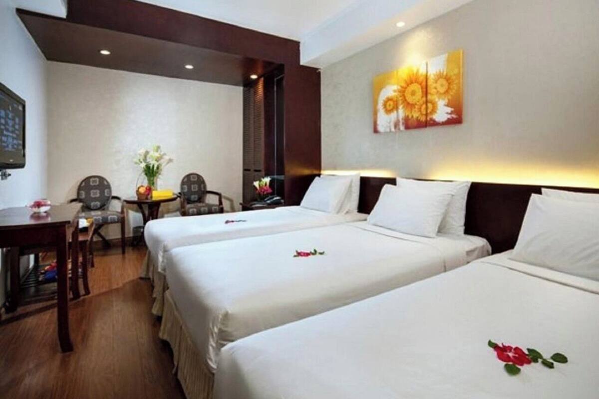 Ein Hotelzimmer mit zwei Betten, einem Schreibtisch und einem Sessel. An der Wand hängt ein Gemälde und auf einem kleinen Tisch stehen Blumen.