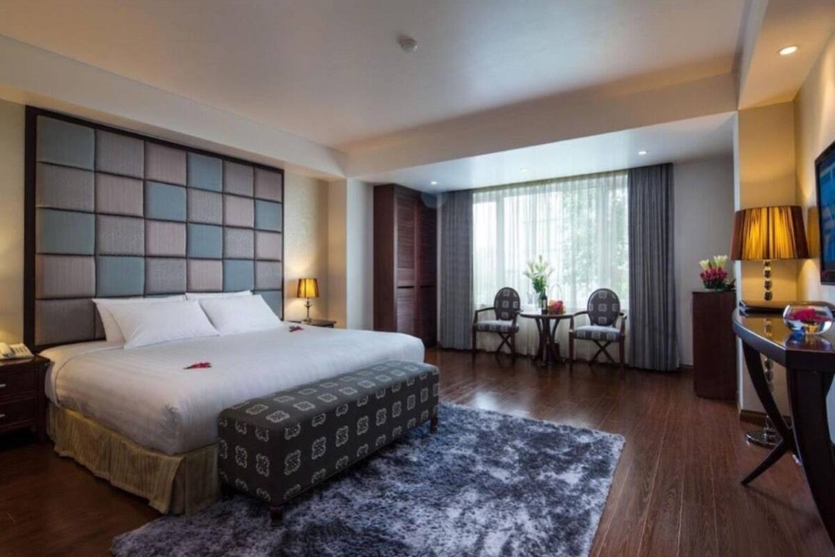 Ein Hotelzimmer mit einem großen Bett, einem Schreibtisch mit Stuhl, einem kleinen Tisch mit Blumen und einem Fernseher an der Wand.