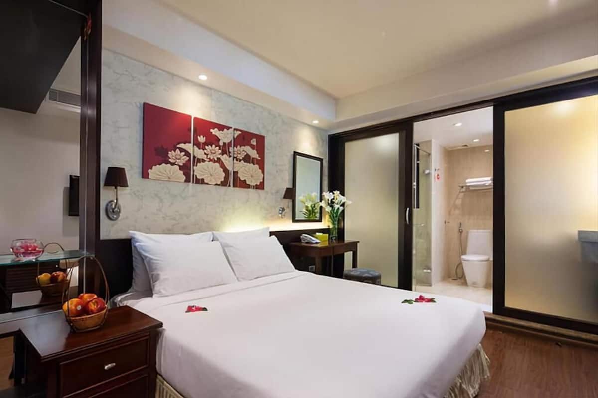 Ein Hotelzimmer mit einem großen Bett, einem Schreibtisch mit Lampe, einer Obstschale und Wandbildern mit floralen Motiven.