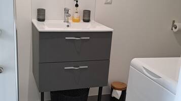 Une salle de bain avec un lavabo blanc, un miroir, un distributeur de serviettes et une poubelle noire.